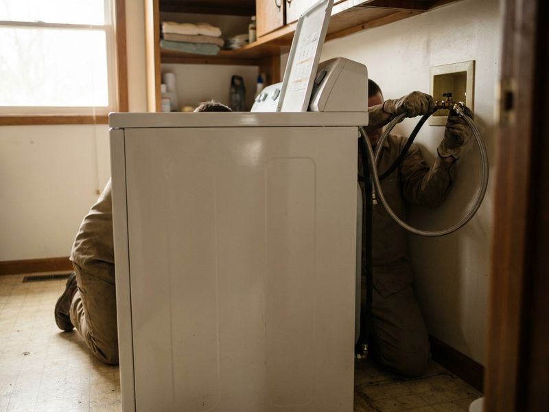 A technician kneeling behind a washing machine, connecting the water supply hose to the wall valves.